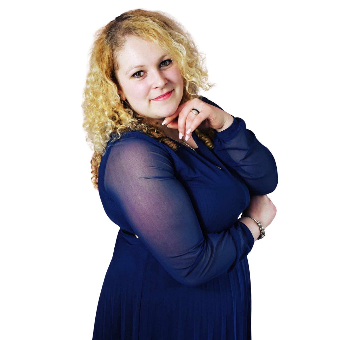 Portret uśmiechniętej kobiety z kręconymi blond włosami, w granatowej sukience z prześwitującymi rękawami, pozującej z dłonią pod brodą na białym tle.