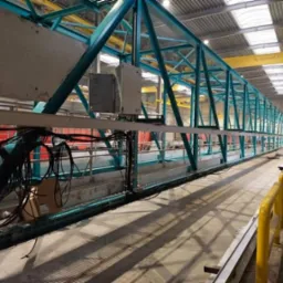 Długa, stalowa konstrukcja kratownicowa pomalowana na niebiesko, z widocznymi spawami, w hali produkcyjnej z żółtym suwnicą w tle.