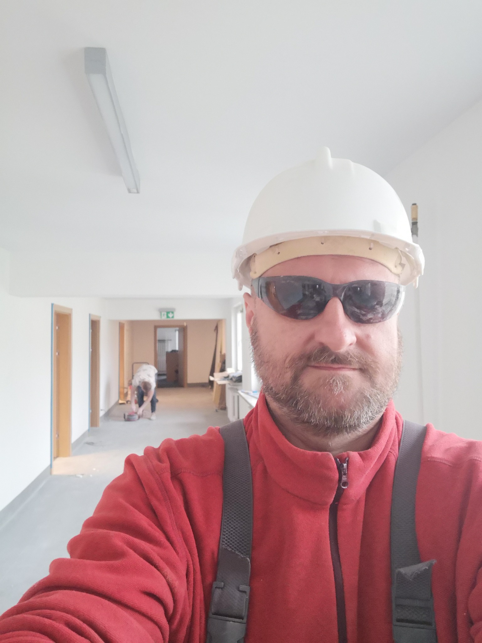 Autoportret pracownika w kasku i okularach ochronnych na tle korytarza w trakcie prac wykończeniowych; w tle widoczna osoba szlifująca podłogę.