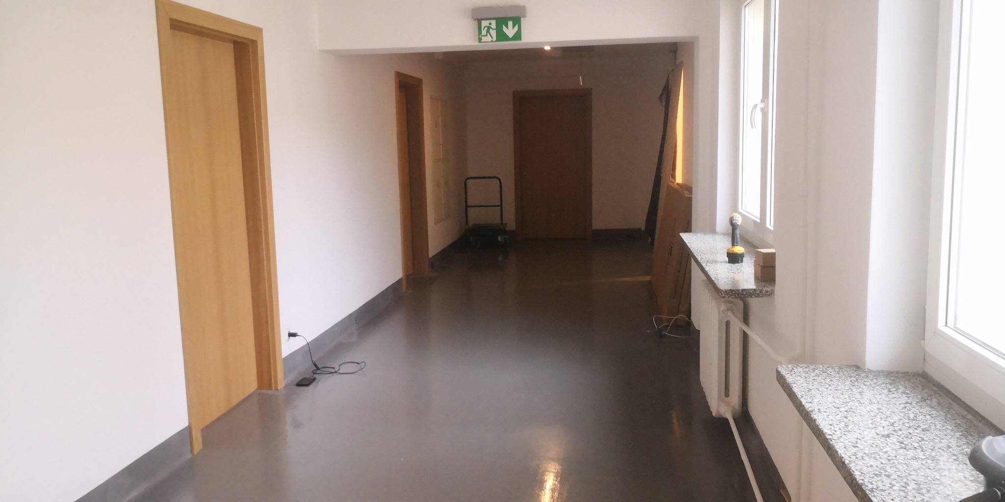 Korytarz po remoncie z nową, błyszczącą posadzką, świeżo pomalowanymi ścianami, drewnianymi drzwiami i widocznym narzędziem na parapecie okiennym.