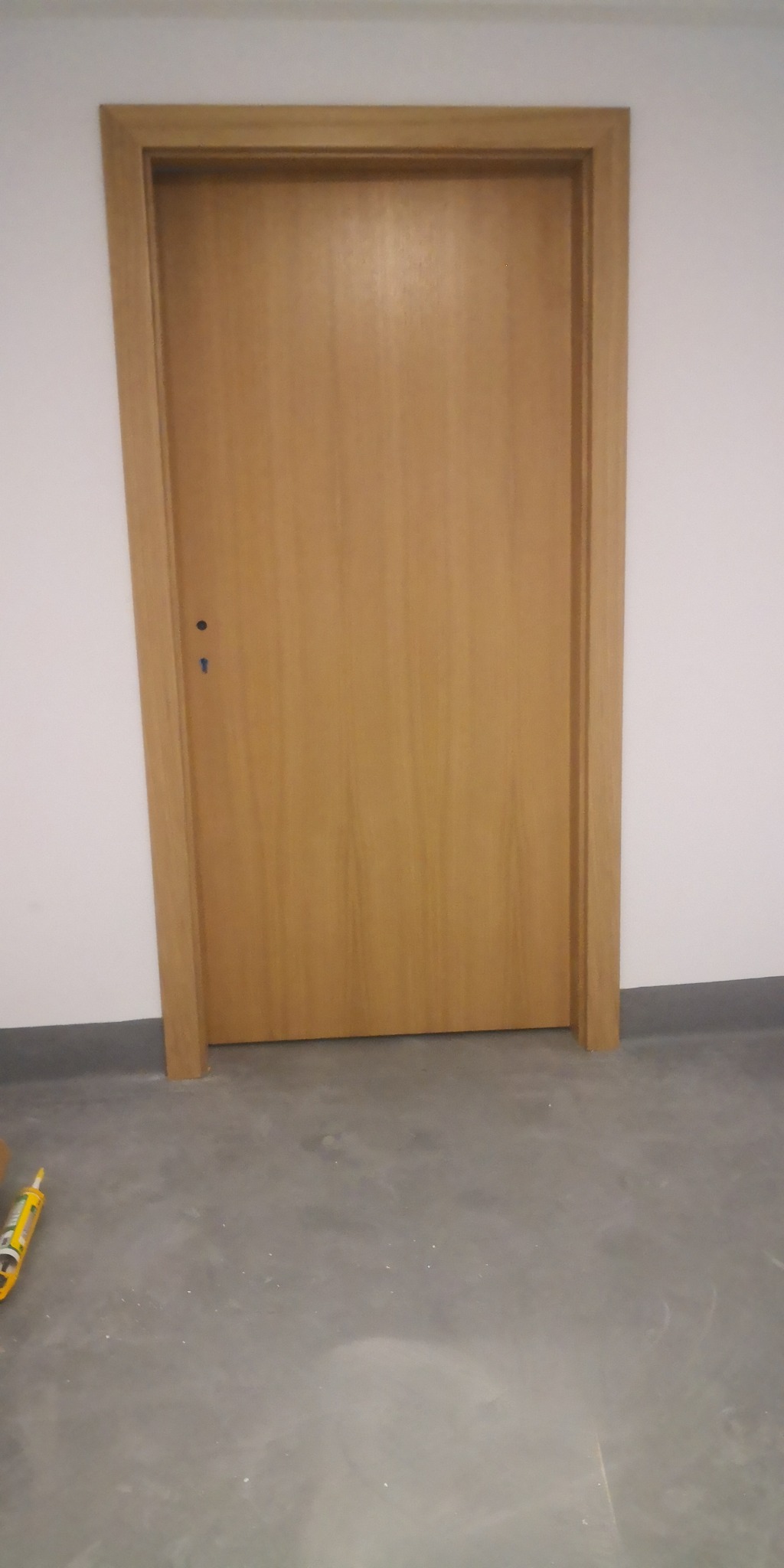 Zamknięte, jasne drewniane drzwi wewnętrzne z ościeżnicą, widoczne na tle białej ściany i szarej podłogi w trakcie montażu, z tubą silikonu na podłodze.