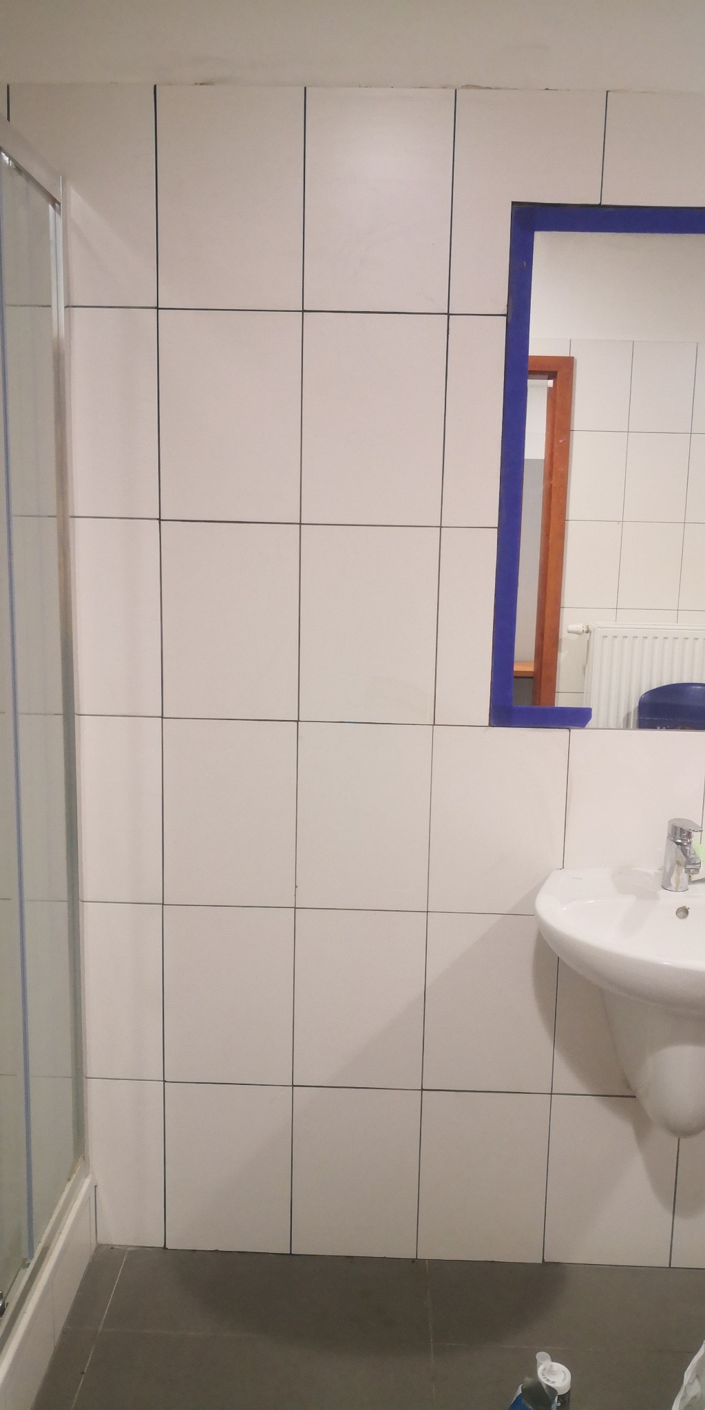 Wykończona łazienka z białymi, kwadratowymi płytkami, narożną kabiną prysznicową i umywalką z baterią. W lustrze odbija się fragment grzejnika i niebieskiego krzesła.