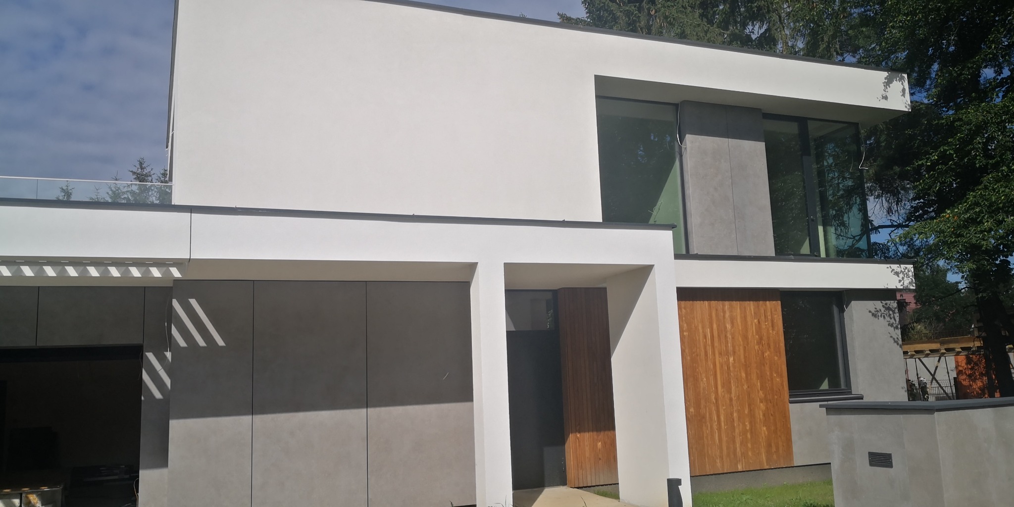 Nowoczesny dom z białą elewacją, szarymi panelami i drewnianymi akcentami, duże okna odbijające zieleń drzew.