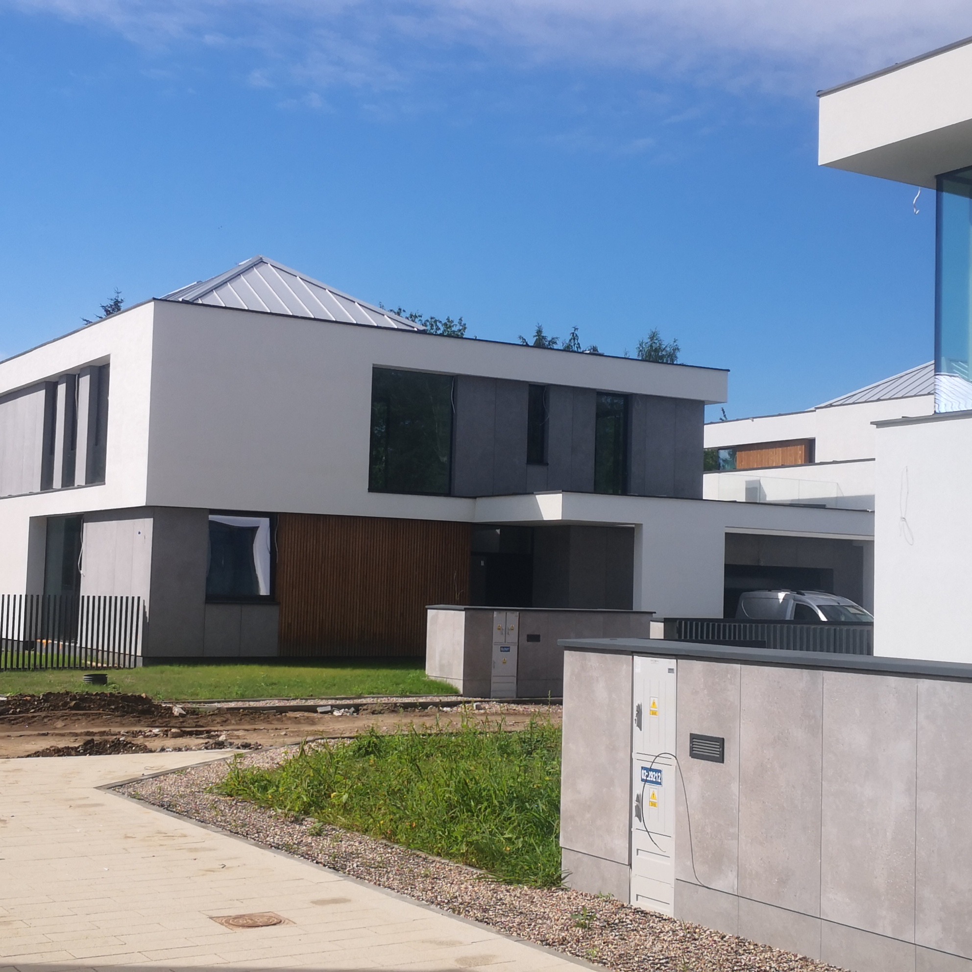 Nowoczesny dom jednorodzinny z płaskim dachem i drewnianą elewacją, widoczne skrzynki elektryczne na ogrodzeniu i teren w trakcie zagospodarowania.