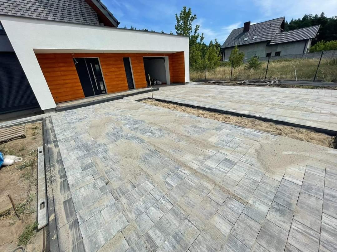 Szara kostka brukowa ułożona przed nowoczesnym domem z drewnianą elewacją, widoczne niedokończone fragmenty z piaskiem i obrzeżami.