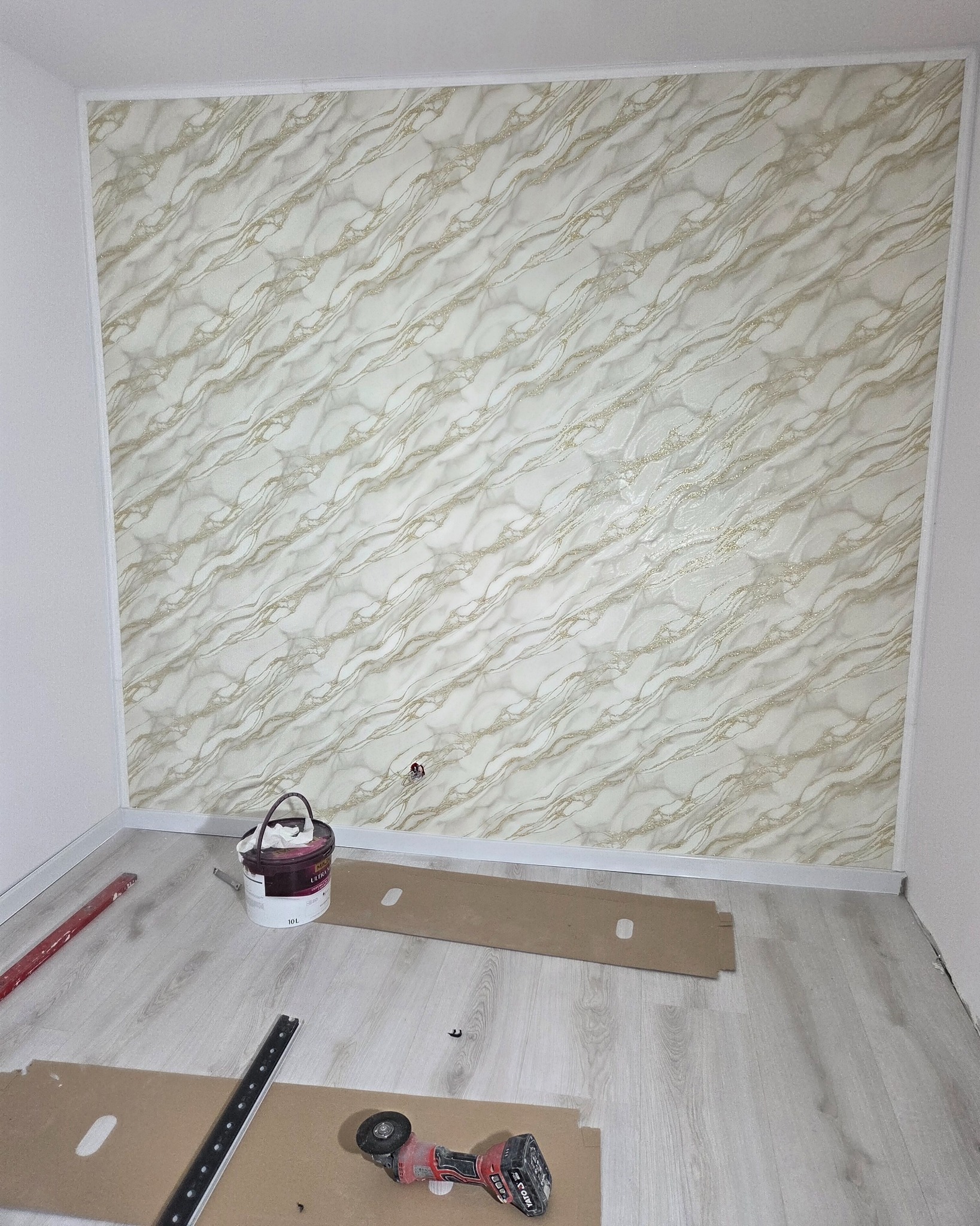 Wykończona ściana z dekoracyjną tapetą w marmurkowy wzór, narzędzia i puszka farby na podłodze. Widok na róg pokoju w trakcie prac wykończeniowych.