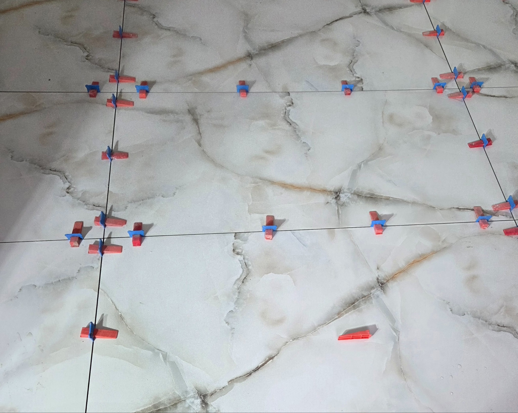Układanie płytek imitujących marmur z użyciem systemu poziomowania. Widoczne krzyżyki dystansowe i kliny. Czarny sznurek wyznacza linie proste.