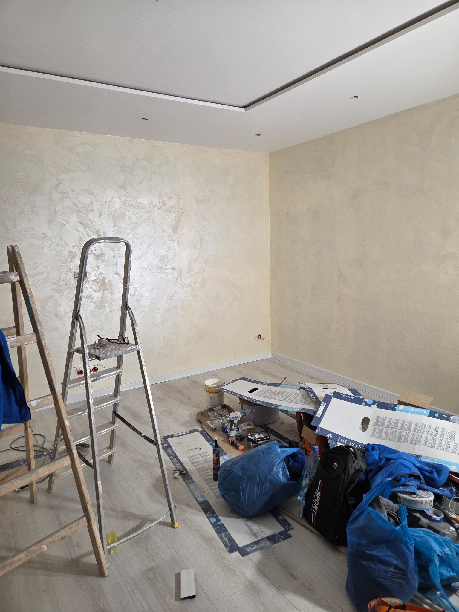 Pokój po gładzi dekoracyjnej w odcieniach beżu, z widocznymi drabinami i narzędziami malarskimi. Widoczne kartony po panelach podłogowych.