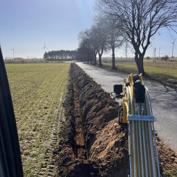 IE24 Piotr Woźniak - Wykopany rów wzdłuż drogi, z widocznym fragmentem koparki. W tle pole, drzewa i turbiny wiatrowe. Prace ziemne związane z instalacją podziemną.
