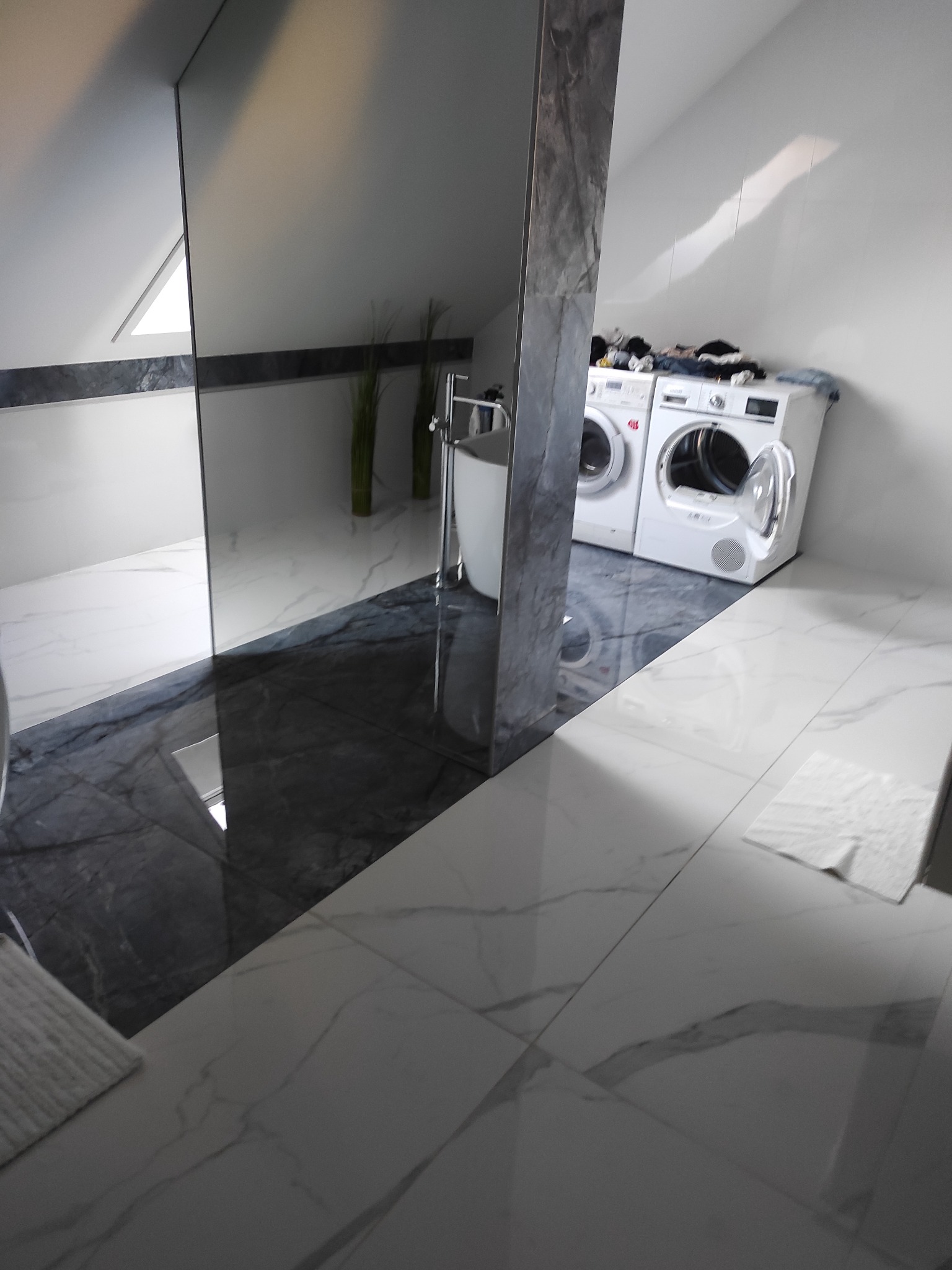 Nowoczesna łazienka na poddaszu z marmurową podłogą i ścianami, pralka i suszarka na tle, widok na okno dachowe.