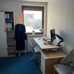 Wnętrze biura: toga adwokacka wisząca na wieszaku, biurko z monitorem i klawiaturą, krzesło biurowe, regał z segregatorami, widok z okna na budynki.