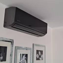 Ciemnoszara klimatyzacja marki Rotenso zamontowana na białej ścianie w pomieszczeniu mieszkalnym, poniżej wiszą trzy zdjęcia w błyszczących, srebrnych ramkach.