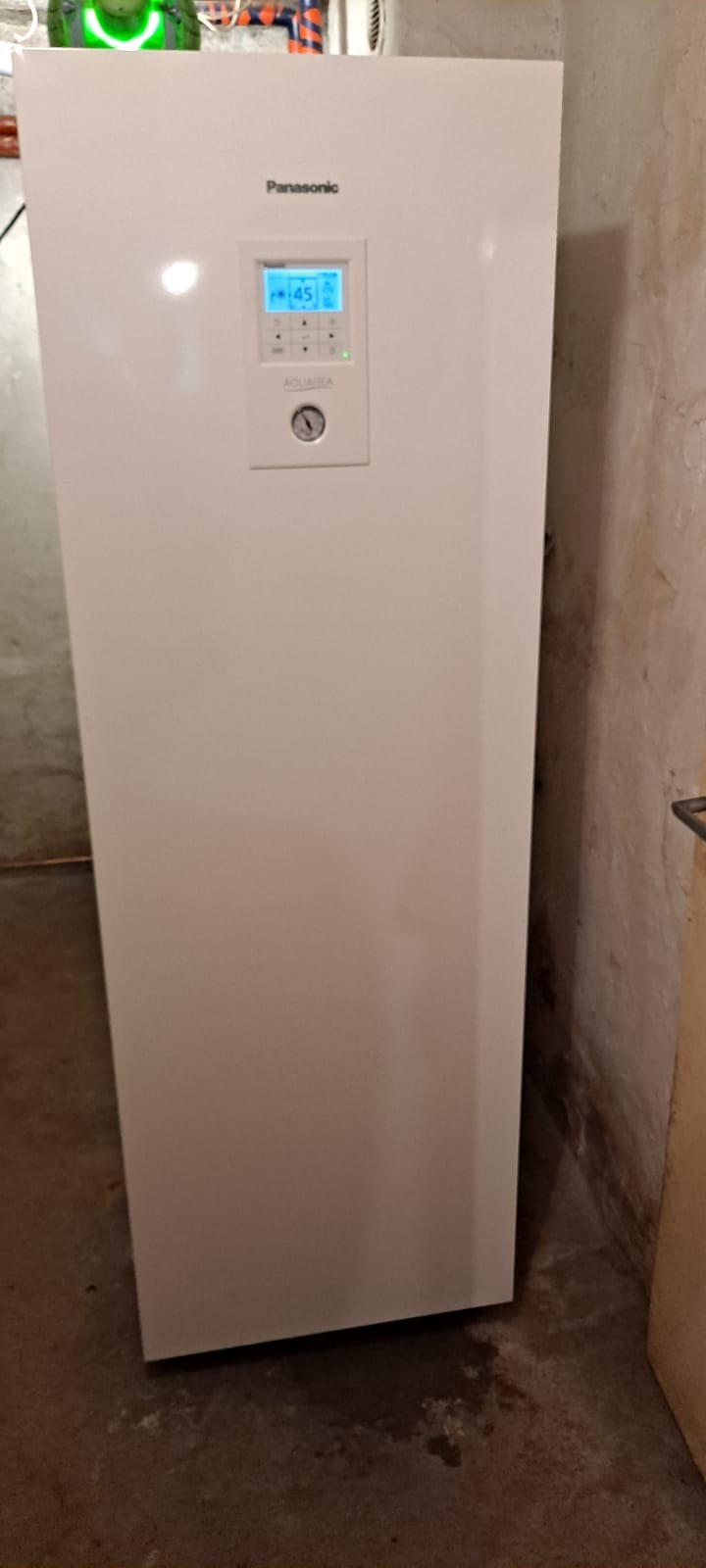 Biała pompa ciepła Panasonic Aquarea z wyświetlaczem pokazującym 45 stopni, zainstalowana w pomieszczeniu z surowymi ścianami i betonową podłogą.
