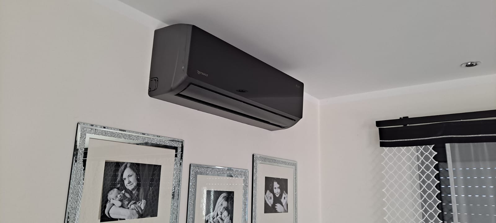 Ciemnoszara klimatyzacja marki Rotenso zamontowana na białej ścianie w pomieszczeniu mieszkalnym, poniżej wiszą trzy zdjęcia w błyszczących, srebrnych ramkach.