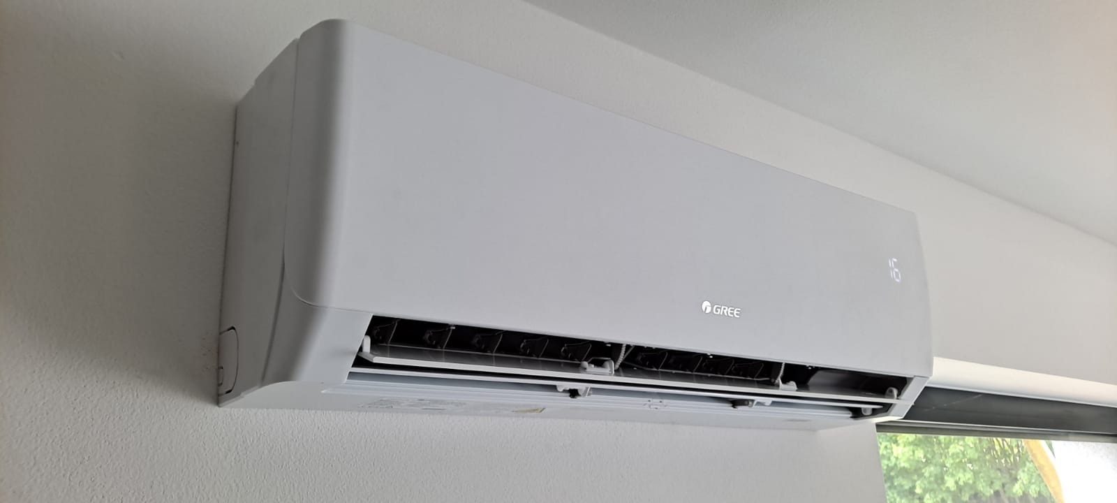 Biała jednostka wewnętrzna klimatyzacji marki Gree zamontowana na ścianie, widoczny wyświetlacz temperatury i otwarte żaluzje.