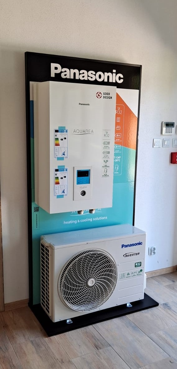 Ekspozycja pompy ciepła Panasonic Aquarea z jednostką wewnętrzną i zewnętrzną na czarnym stojaku z informacjami o produkcie, prezentowana w salonie sprzedaży.