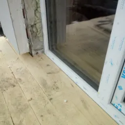 Montaż okna z widoczną pianką montażową, ociepleniem ściany i surową drewnianą podłogą, widoczne detale profilu okiennego VEKA.