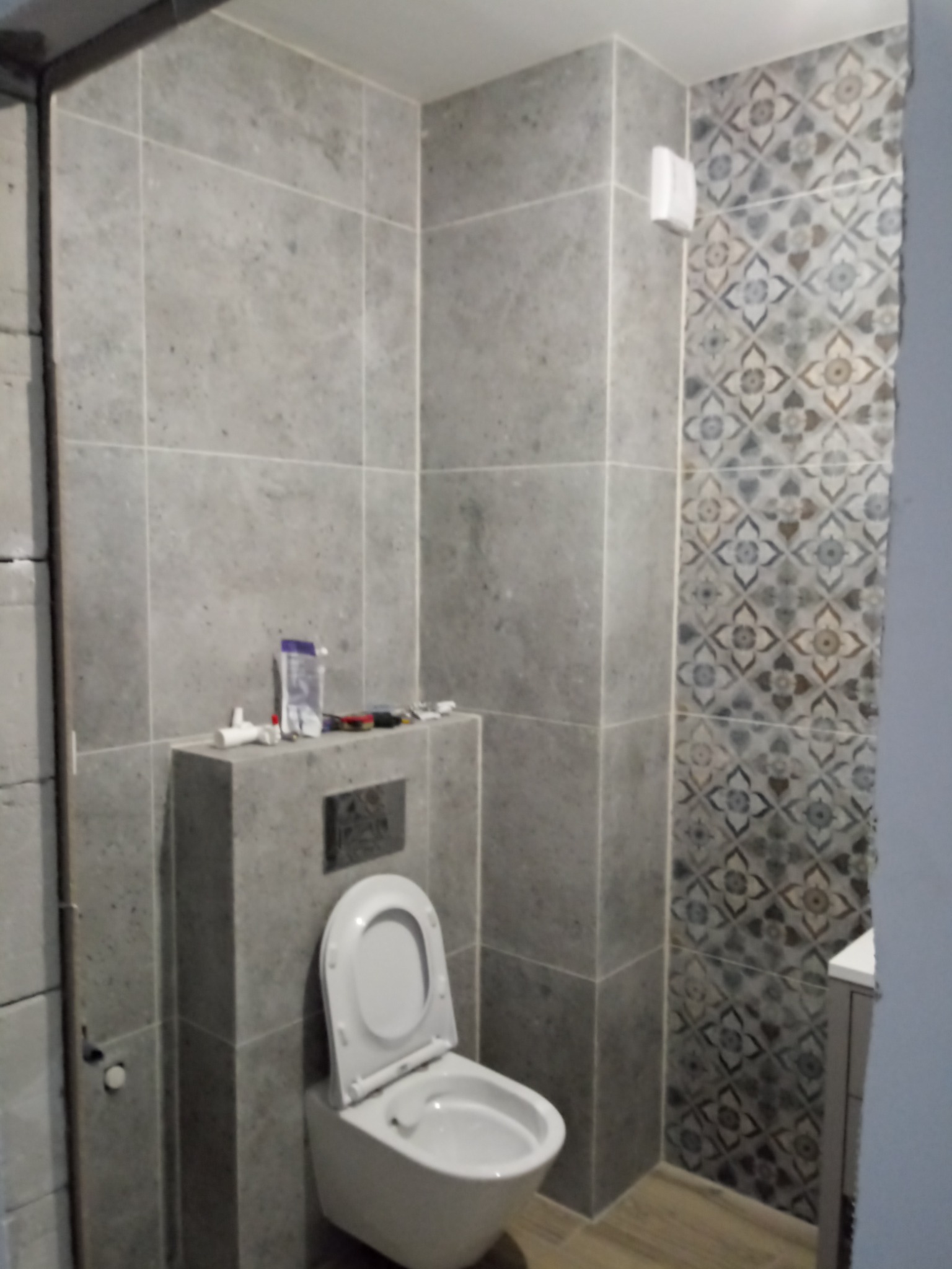 Szara łazienka z toaletą podwieszaną na stelażu obudowanym płytkami, widoczne narzędzia i akcesoria hydrauliczne na obudowie stelaża, ściana wykończona płytkami w dwa wzory.