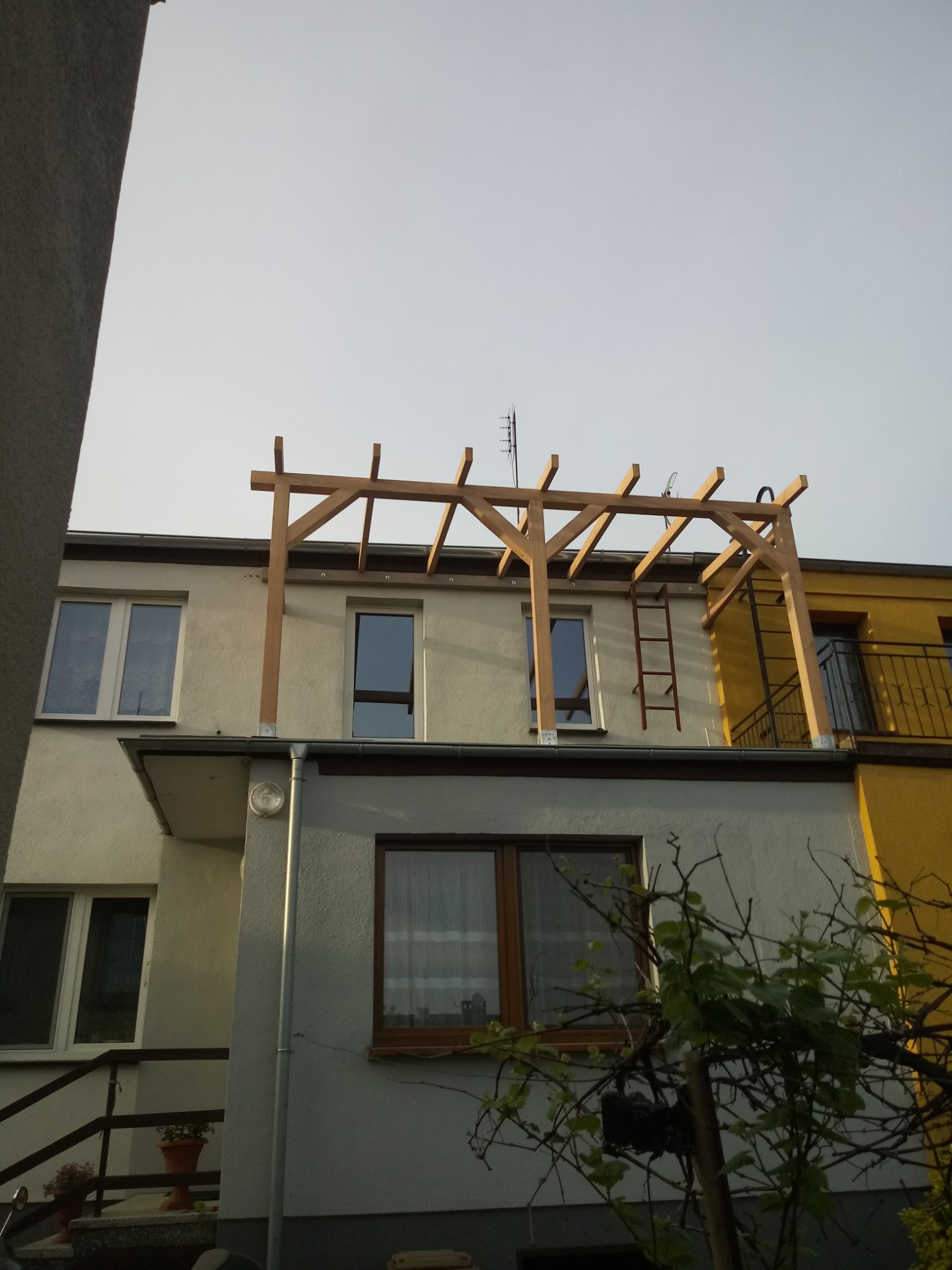 Montaż drewnianej konstrukcji, prawdopodobnie pergoli, na dachu dwupiętrowego budynku, widoczna drabina oparta o ścianę, jasna elewacja budynku.