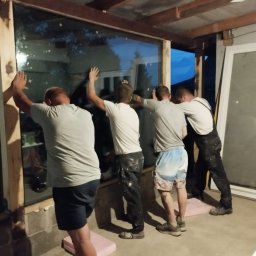 Robert K - Czterech mężczyzn instaluje duże okno w surowym otworze okiennym, podpierając je piankowymi blokami. Jeden z nich opiera się całym ciałem o okno, aby je utrzymać w miejscu.