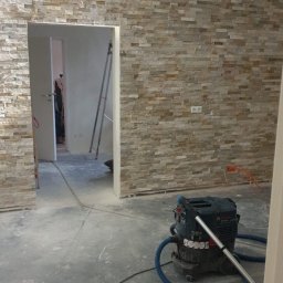 Robert K - Ściana wyłożona dekoracyjną okładziną kamienną w trakcie prac, widoczny odkurzacz przemysłowy i wiadro z zaprawą, surowy beton na podłodze, w tle metalowa drabina.