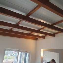 Robert K - Biały sufit panelowy z drewnianymi belkami konstrukcyjnymi w jasnym wnętrzu domu z widokiem na zewnątrz przez przesuwne drzwi balkonowe.