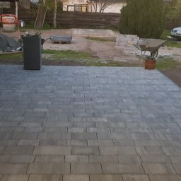 Robert K - Ułożona szara kostka brukowa na podwórku, widoczne donice, taczka, samochód i materiały budowlane w tle.