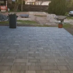 Ułożona szara kostka brukowa na podwórku, widoczne donice, taczka, samochód i materiały budowlane w tle.