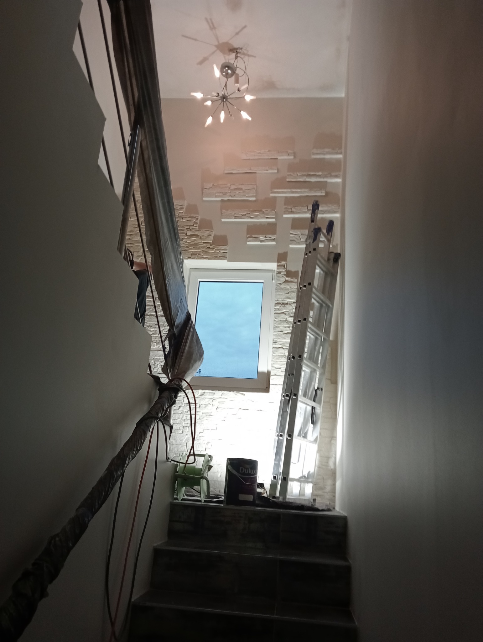 Klatka schodowa w trakcie prac, ściana z dekoracyjną okładziną imitującą kamień, aluminiowa drabina oparta o ścianę, wiadro z farbą Dulux na schodach, poręcz zabezpieczona folią malarską.