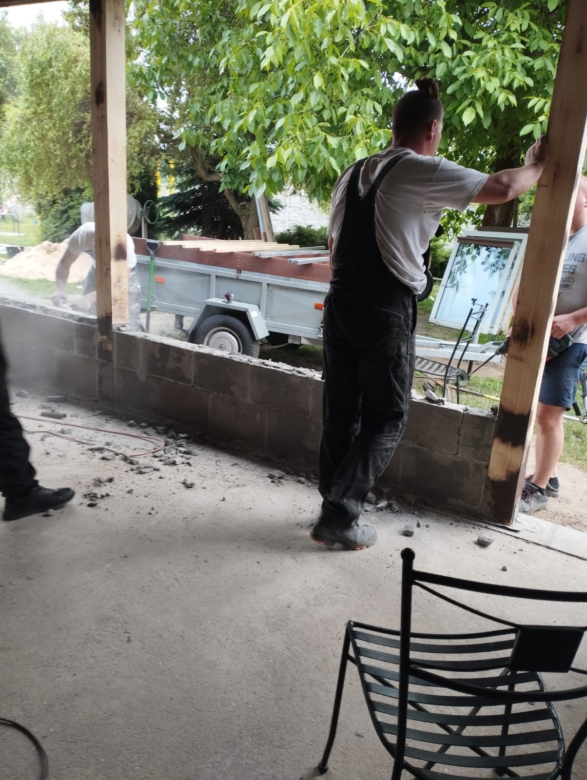 Pracownik w kombinezonie opiera się o drewnianą ramę podczas prac murarskich, w tle przyczepa z drewnem i okno oprawione w ramę