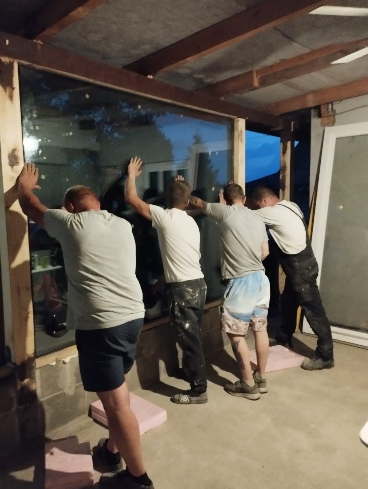 Czterech mężczyzn instaluje duże okno w surowym otworze okiennym, podpierając je piankowymi blokami. Jeden z nich opiera się całym ciałem o okno, aby je utrzymać w miejscu.