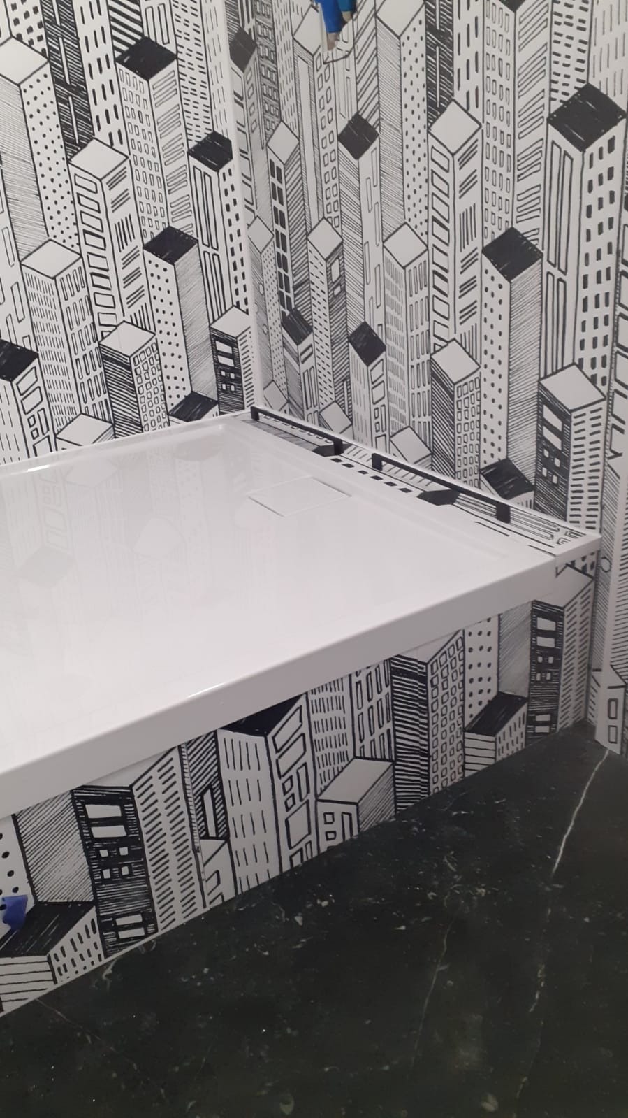 Biały brodzik prysznicowy z niskim progiem, zamontowany na tle ściany z tapetą przedstawiającą czarno-biały rysunek miejskiego krajobrazu.