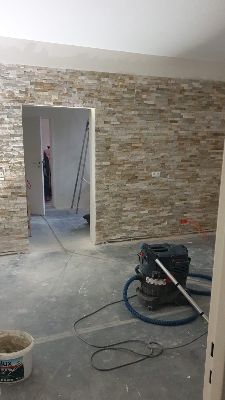 Ściana wyłożona dekoracyjną okładziną kamienną w trakcie prac, widoczny odkurzacz przemysłowy i wiadro z zaprawą, surowy beton na podłodze, w tle metalowa drabina.