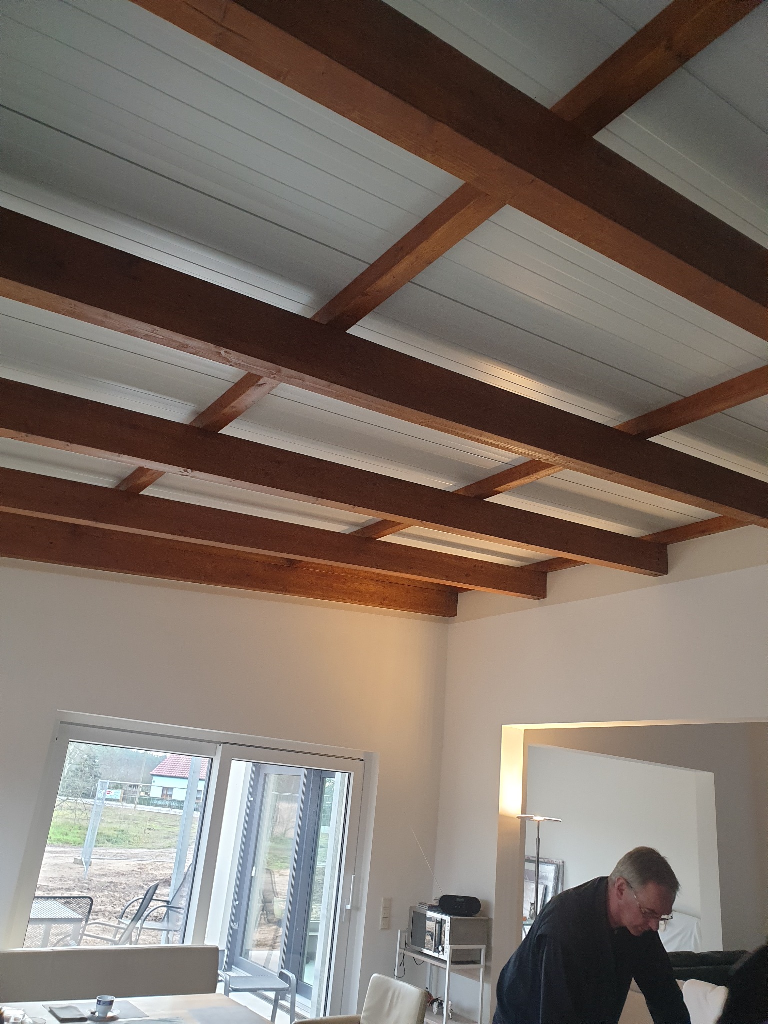 Biały sufit panelowy z drewnianymi belkami konstrukcyjnymi w jasnym wnętrzu domu z widokiem na zewnątrz przez przesuwne drzwi balkonowe.