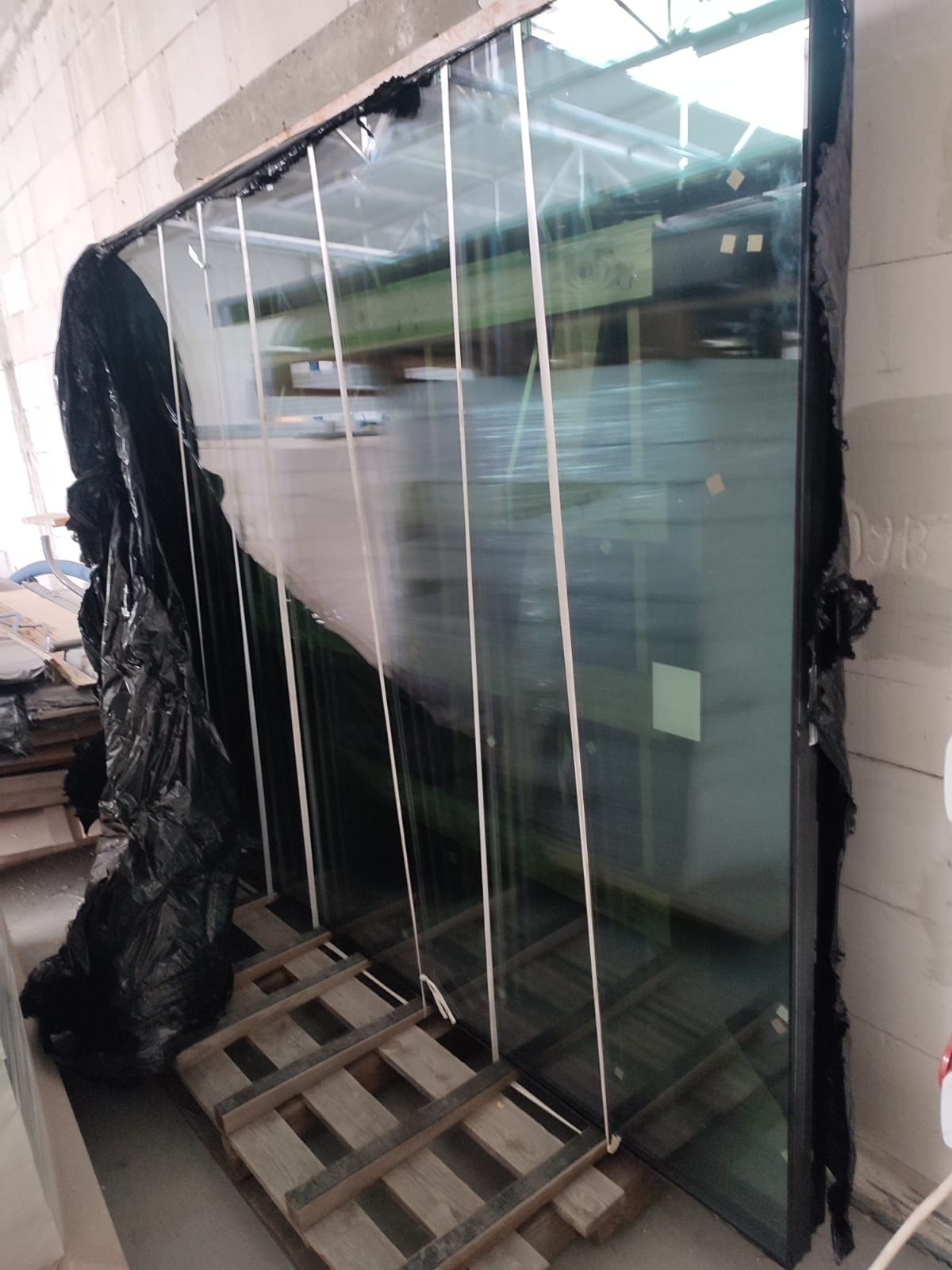 Duże okno balkonowe w czarnej ramie, zabezpieczone folią, oparte na palecie w surowym wnętrzu.