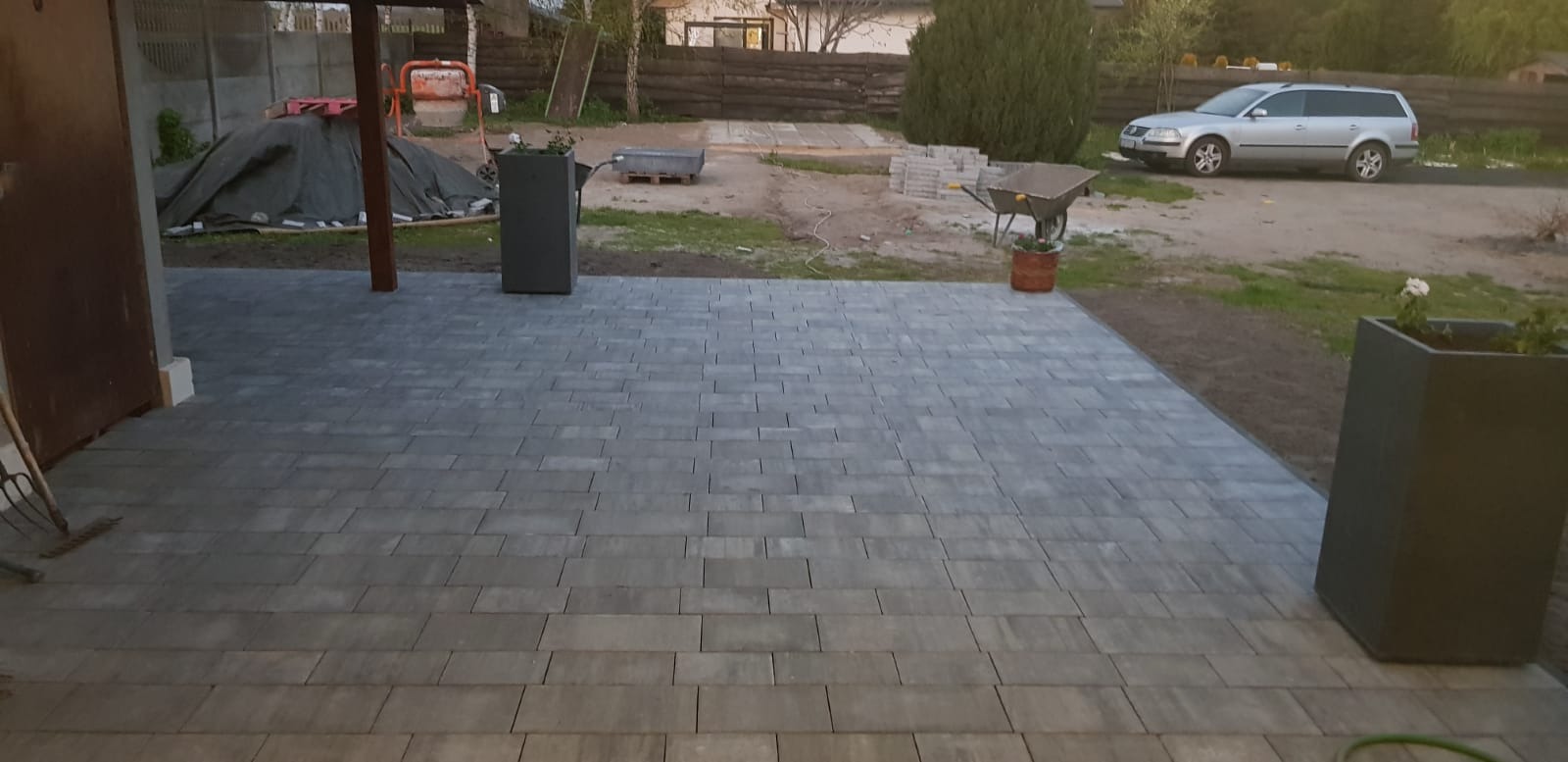Ułożona szara kostka brukowa na podwórku, widoczne donice, taczka, samochód i materiały budowlane w tle.
