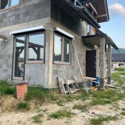 Icon - Surowy stan budynku z szarego betonu komórkowego, z zamontowanymi roletami zewnętrznymi w kolorze srebrnym nad oknami, widoczne rusztowania i narzędzia budowlane na zewnątrz.