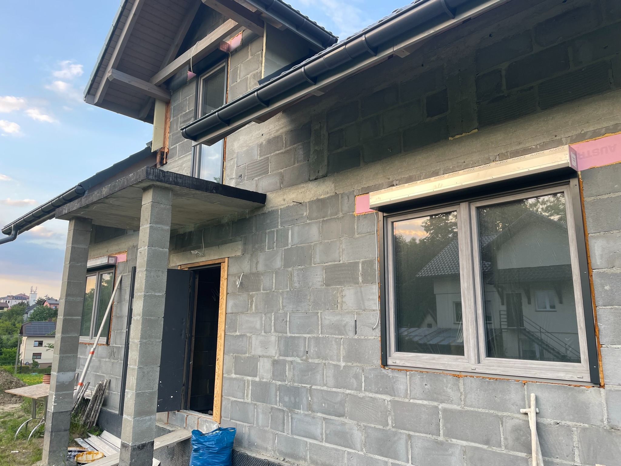 Surowy stan elewacji domu w trakcie montażu rolet podtynkowych w kolorze antracytowym, widoczne okna z zamontowanymi prowadnicami i częściowo schowanymi kasetami rolet, oraz fragmenty ocieplenia.
