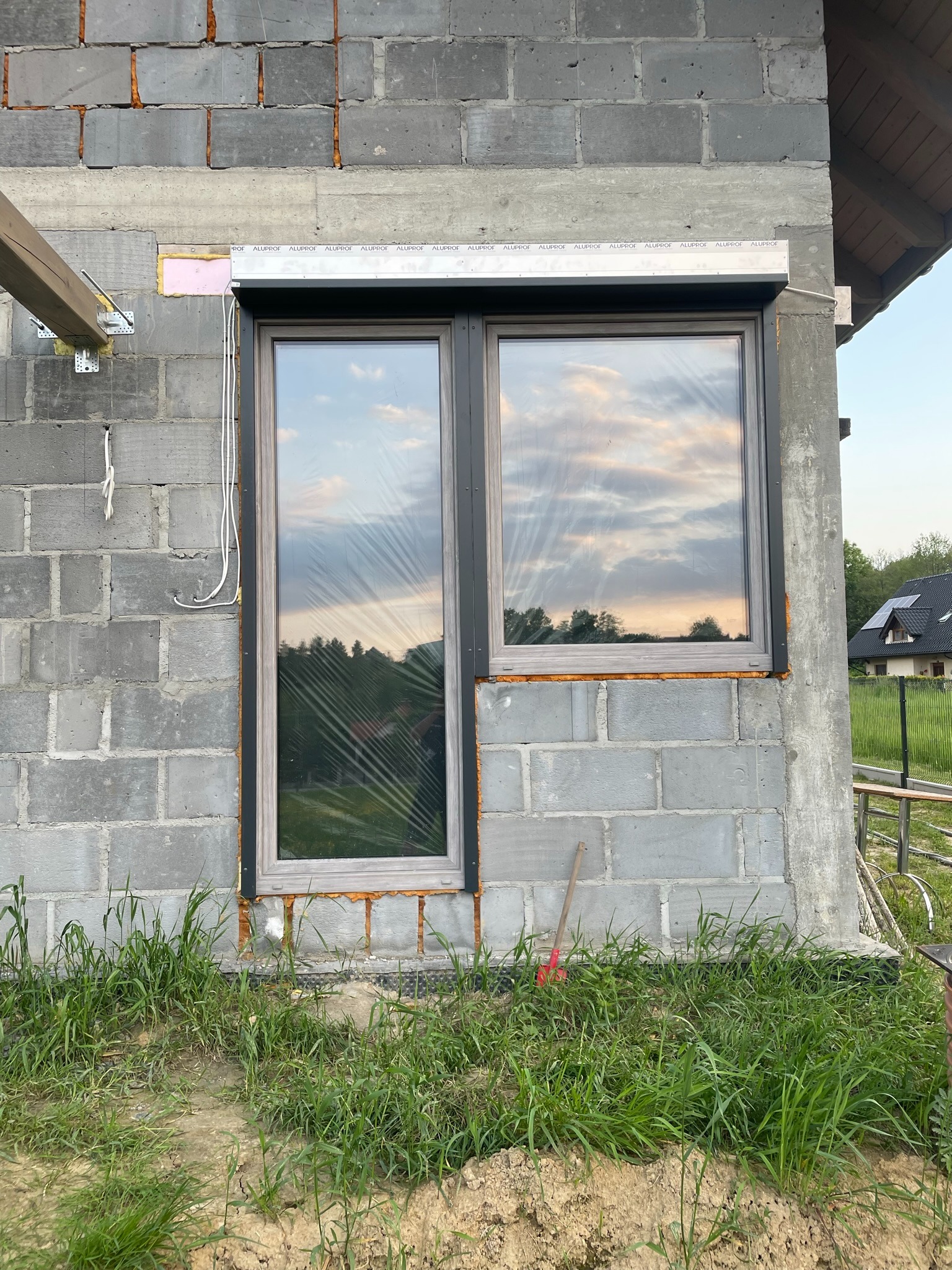 Montaż rolet podtynkowych w kolorze antracyt na oknie w surowym budynku z bloczków w Kobiernicach, widoczne przewody elektryczne i trawa przed budynkiem.