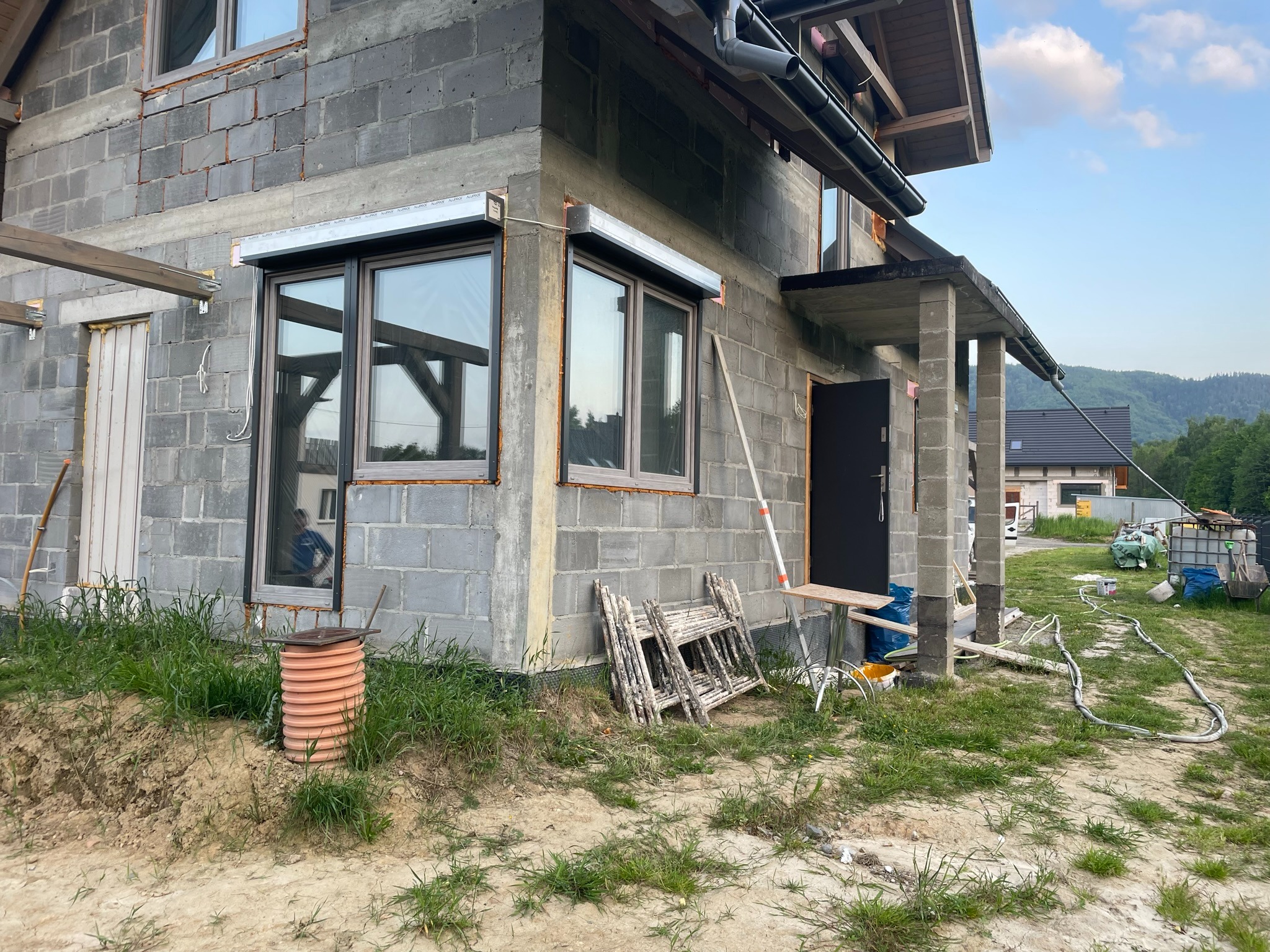 Surowy stan budynku z szarego betonu komórkowego, z zamontowanymi roletami zewnętrznymi w kolorze srebrnym nad oknami, widoczne rusztowania i narzędzia budowlane na zewnątrz.