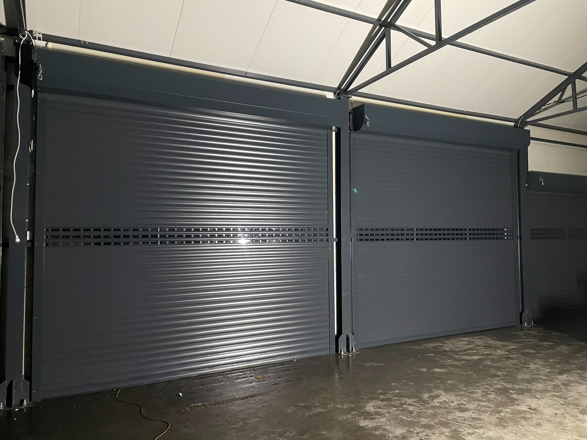 Dwie antracytowe bramy rolowane z profilem PA77, widoczne w garażu pod metalową konstrukcją dachu, odbijające światło na mokrej posadzce.