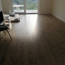 Marcin Łuczak - Ułożone panele podłogowe w jasnym pomieszczeniu z dużym oknem balkonowym i widokiem na zieleń, widoczne opakowania po panelach i krzesło.