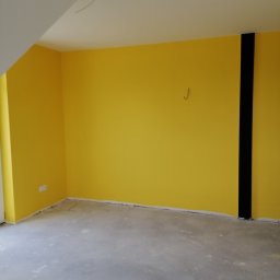 Marcin Łuczak - Świeżo pomalowane na żółto ściany pokoju w Łodzi z widocznym czarnym pasem na jednej ze ścian oraz wystającymi kablami z sufitu, przygotowane do dalszych prac wykończeniowych.