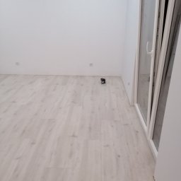 Marcin Łuczak - Jasne panele podłogowe ułożone w pustym, pomalowanym na biało pokoju, z widocznym poziomnicą laserową przy ścianie i oknem balkonowym.