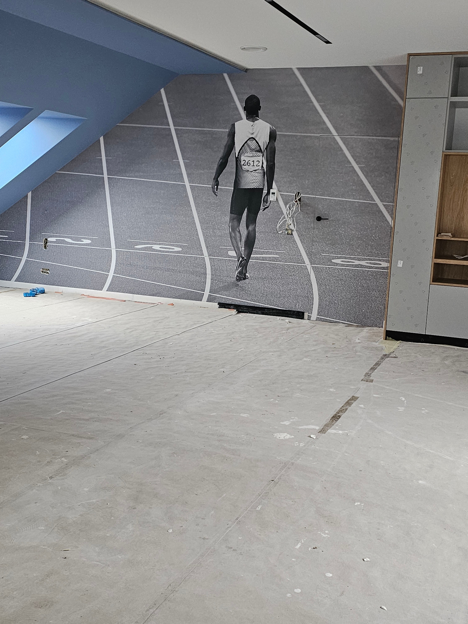 Surowy betonowy podkład podłogowy w pomieszczeniu z niebieskim sufitem i fototapetą przedstawiającą bieżnię lekkoatletyczną z biegnącym sportowcem w tle, obok zabudowa meblowa.