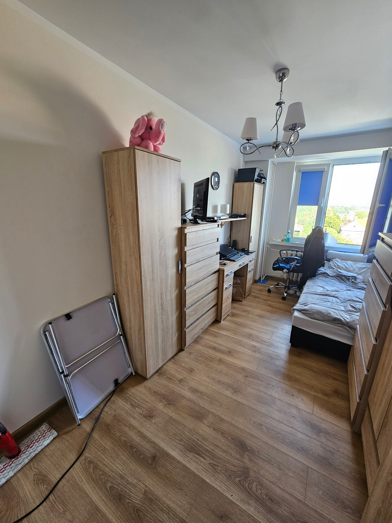 Pokój młodzieżowy z drewnianymi meblami, biurkiem z komputerem i łóżkiem przy oknie. Na szafie pluszowy różowy słoń. Na podłodze panele i składane krzesło.