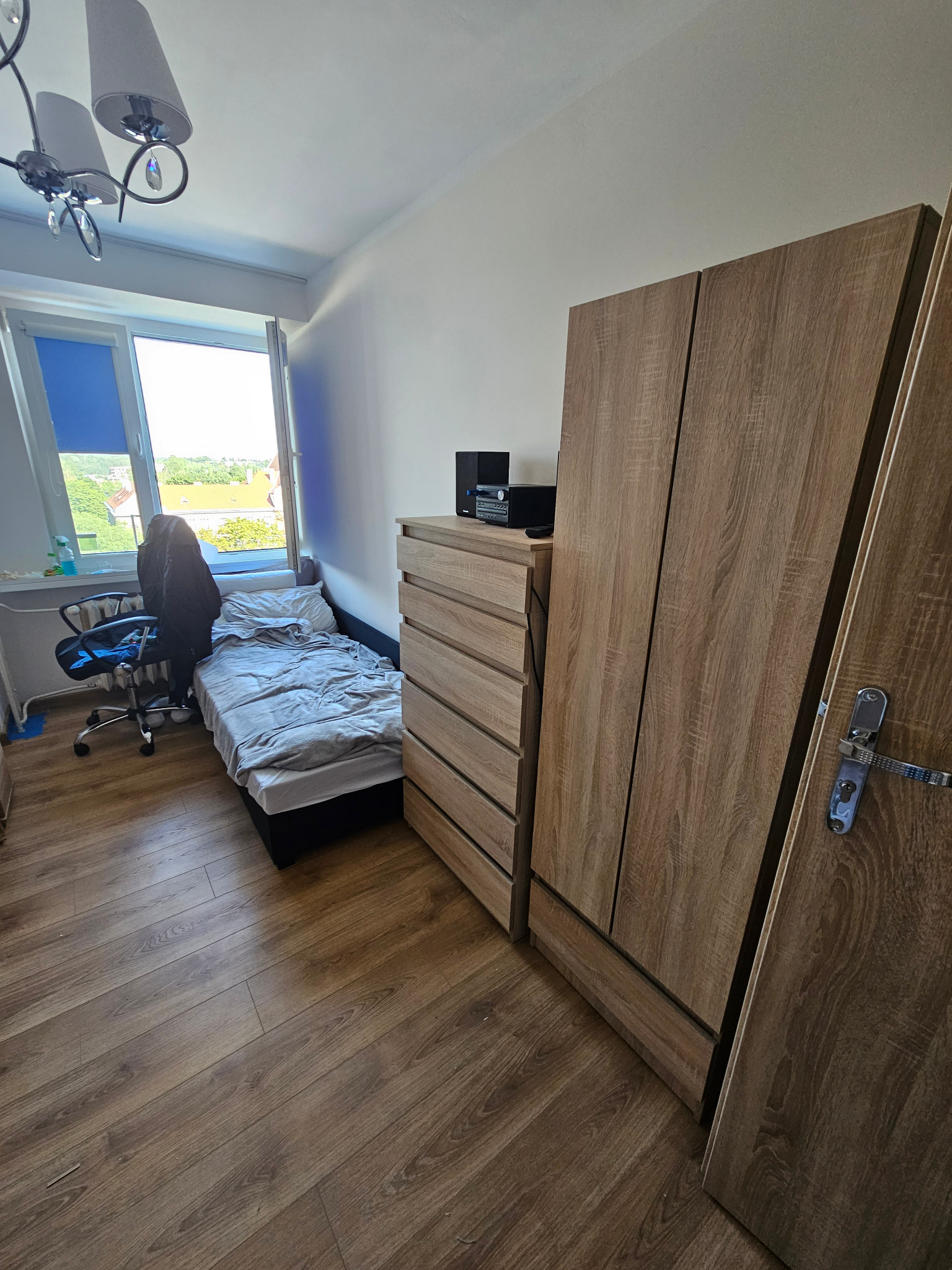 Pokój z łóżkiem, komodą i szafą w odcieniach brązu, widok na zewnątrz przez okno z niebieską roletą, na podłodze panele imitujące drewno.