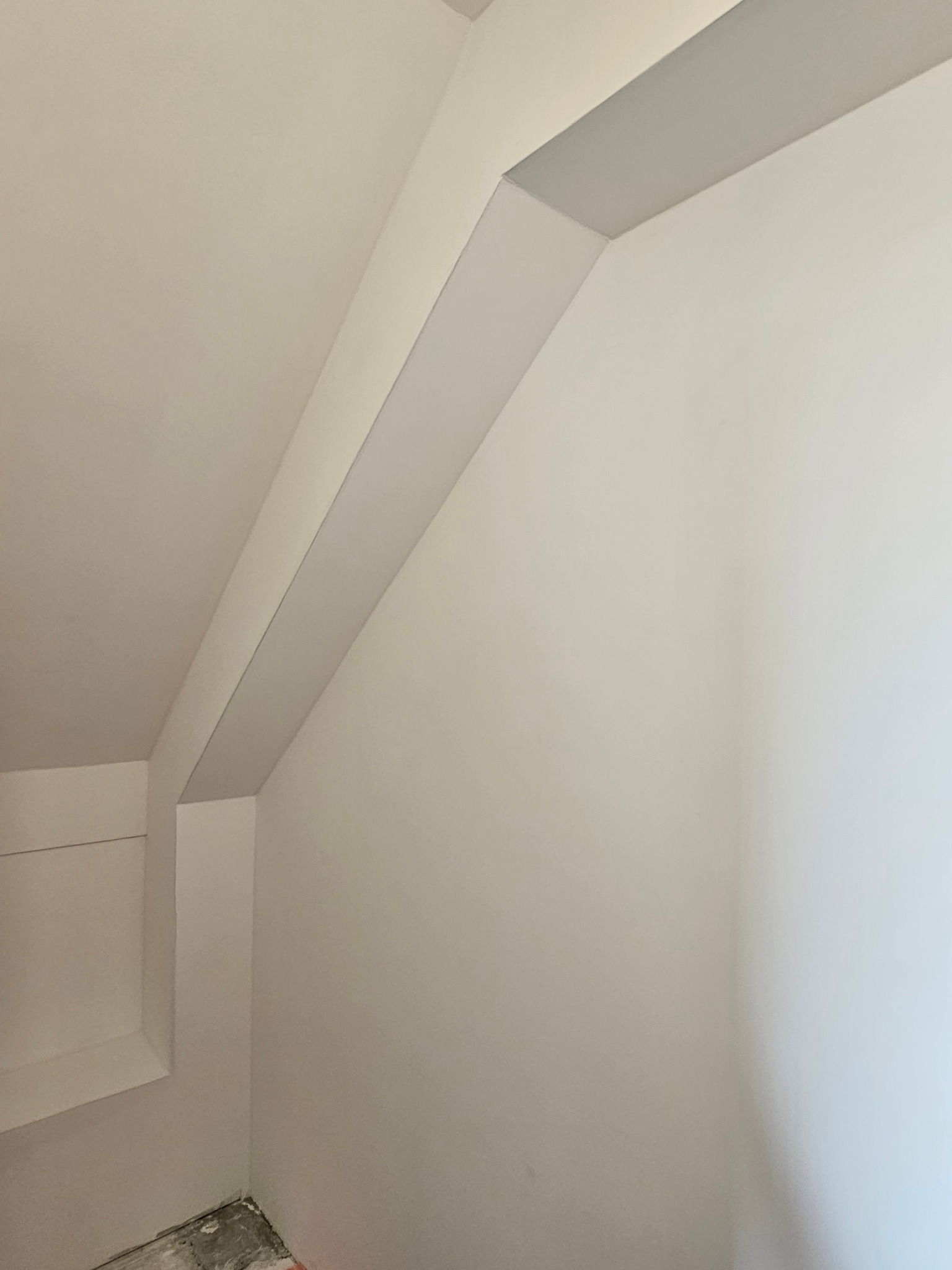 Wnętrze poddasza po malowaniu na biało, widoczna skośna ściana z wystającym elementem konstrukcyjnym i wnęką, surowa podłoga w rogu.