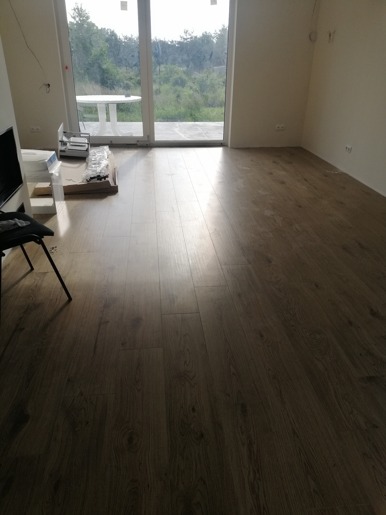 Ułożone panele podłogowe w jasnym pomieszczeniu z dużym oknem balkonowym i widokiem na zieleń, widoczne opakowania po panelach i krzesło.