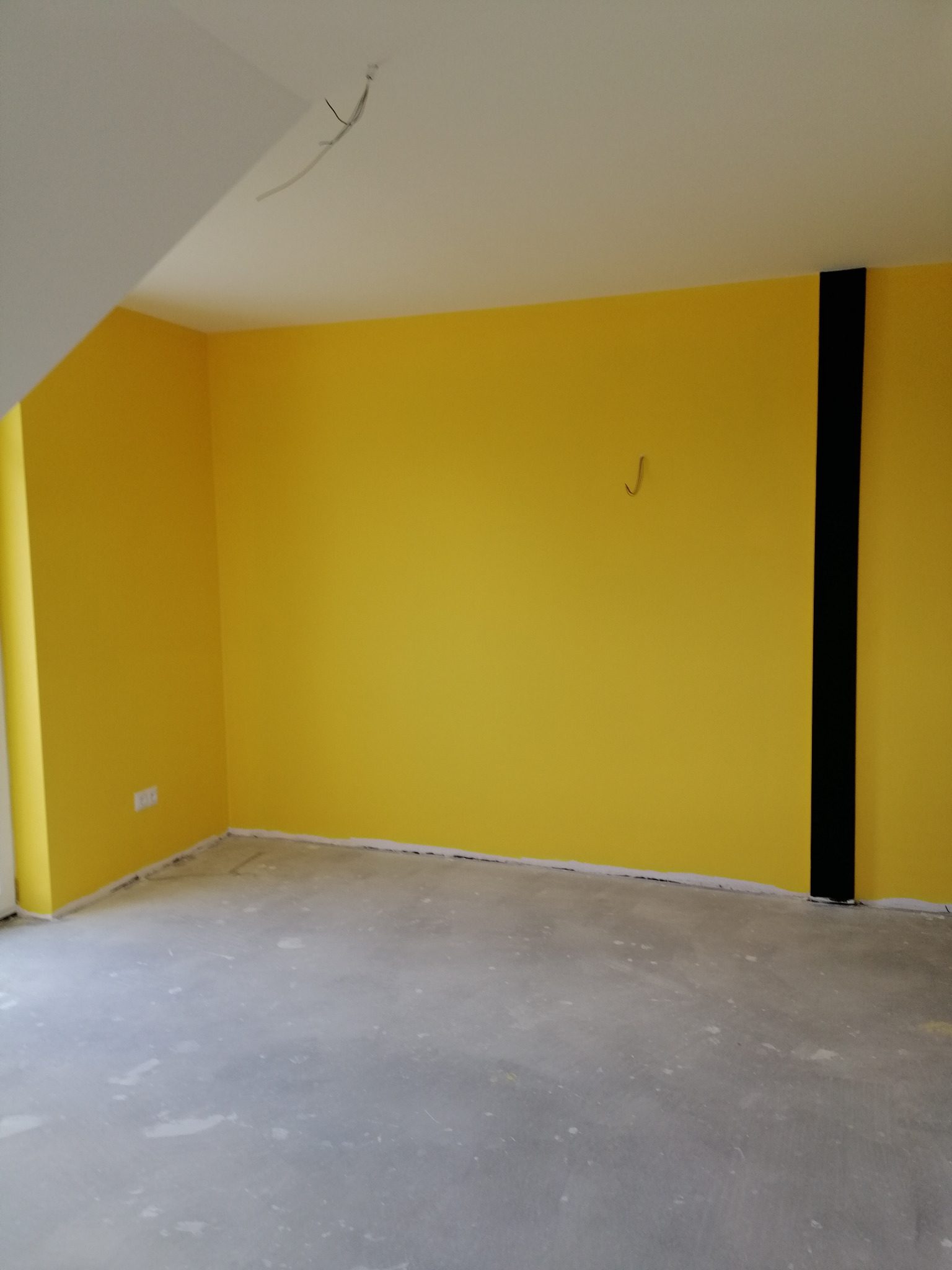 Świeżo pomalowane na żółto ściany pokoju w Łodzi z widocznym czarnym pasem na jednej ze ścian oraz wystającymi kablami z sufitu, przygotowane do dalszych prac wykończeniowych.